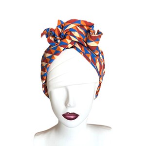 May include: A headwrap featuring a geometric pattern of triangles in shades of orange, blue, and cream. The fabric is tied at the top in a knotted style, covering the crown of a white mannequin head with dark lips, showcasing a vibrant textile design.