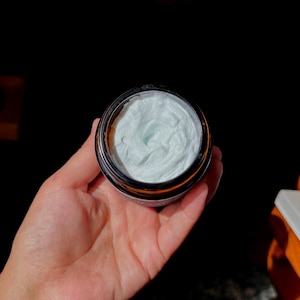 Op de afbeelding: Een potje lichtblauwe crème met een donkerbruine rand en een zilverkleurig deksel. Het potje wordt in een hand gehouden tegen een donkere achtergrond. De crème heeft een gladde, getextureerde uitstraling.