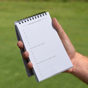 May include: A white notepad with a black spiral binding. The notepad has the text "Mid Round Notes" at the top and the questions "What feels good today?" and "What doesn't feel good today?" written on it.