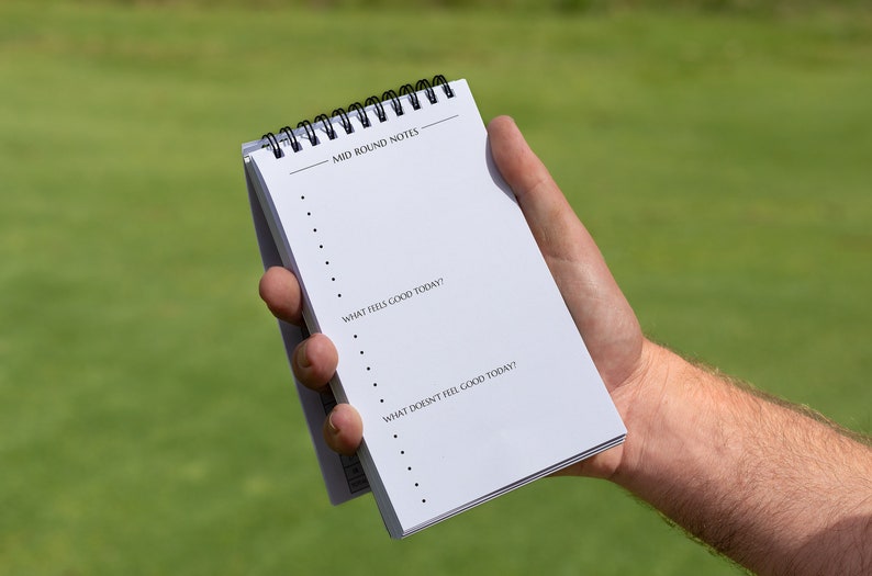 May include: A white notepad with a black spiral binding. The notepad has the text "Mid Round Notes" at the top and the questions "What feels good today?" and "What doesn't feel good today?" written on it.