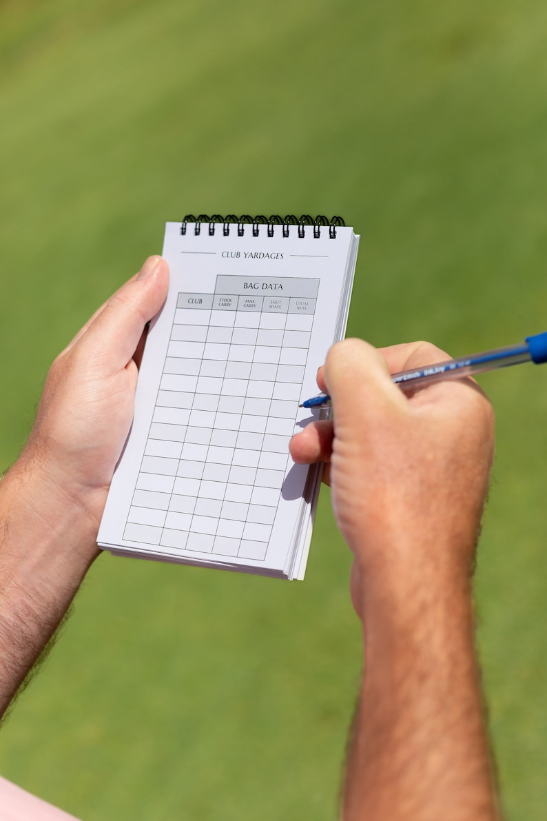 May include: A white spiral bound notebook with the title "Club Yardages" and a table labelled "Bag Data" with columns for "Club", "Stock Carry", "Max Carry", "Shot Shape", and "Usual Yardage".