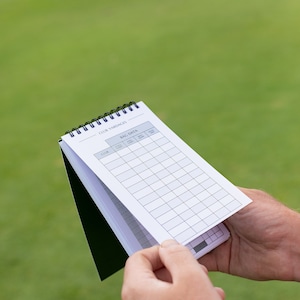 May include: A white spiral bound notebook with the words "Club Yardages" and "Bag Data" printed on the cover. The notebook has columns for "Club", "Loft", "Stock Shaft", "Max Carry", "Shot Shape", and "Club Yardage".