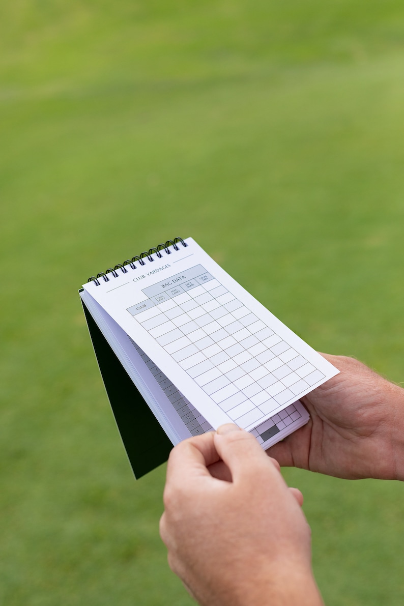May include: A white spiral bound notebook with the words "Club Yardages" and "Bag Data" printed on the cover. The notebook has columns for "Club", "Loft", "Stock Shaft", "Max Carry", "Shot Shape", and "Club Yardage".