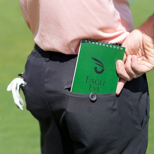 May include: A green spiral bound notebook with the text "EAGLE EYE" and a stylised eye logo. The notebook is in a back pocket of black trousers.