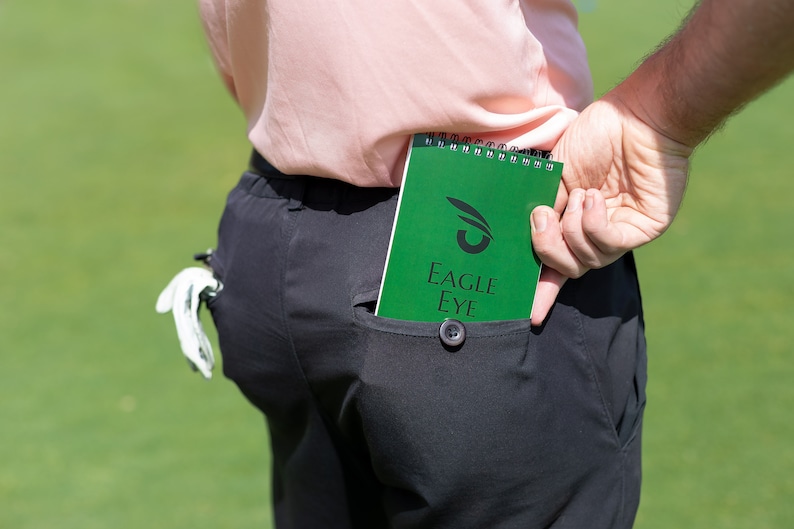 May include: A green spiral bound notebook with the text "EAGLE EYE" and a stylised eye logo. The notebook is in a back pocket of black trousers.