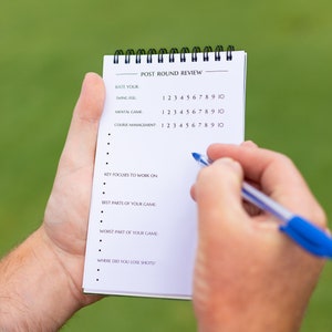 May include: A white notepad with a black spiral binding. The notepad has the title "Post Round Review" and a list of questions for a golfer to rate their performance. The questions include "Rate Your Swing Feel", "Mental Game", and "Course Management".