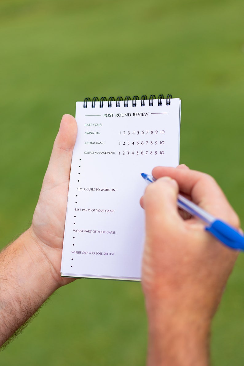 May include: A white notepad with a black spiral binding. The notepad has the title "Post Round Review" and a list of questions for a golfer to rate their performance. The questions include "Rate Your Swing Feel", "Mental Game", and "Course Management".