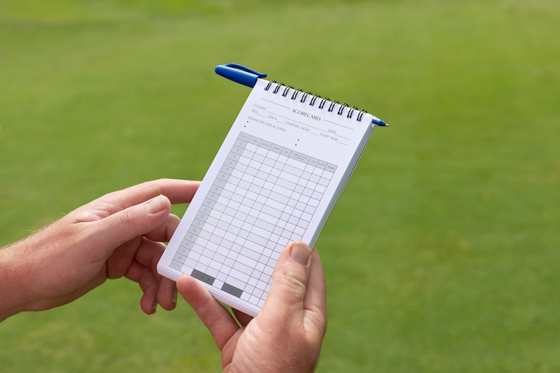 May include: A white golf scorecard with a blue pen on top. The scorecard has columns for each hole, with spaces for the player's score, putts, and other information.