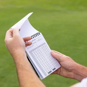 May include: A white golf scorecard with a black spiral binding. The scorecard is filled with columns and rows for recording scores.