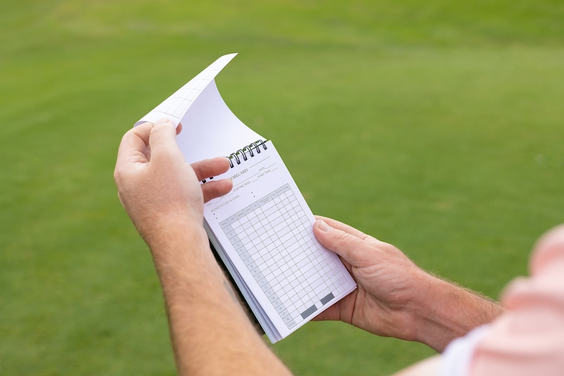 May include: A white golf scorecard with a black spiral binding. The scorecard is filled with columns and rows for recording scores.