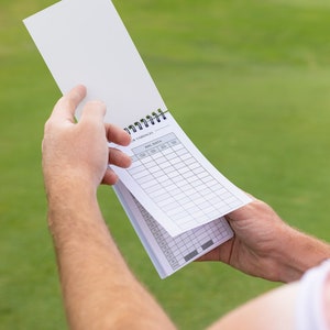 May include: A white golf scorecard with a black spiral binding. The scorecard has columns for each hole, with headings for "Yardages", "Par", "Score", "Putts", "Penalties", and "Total".
