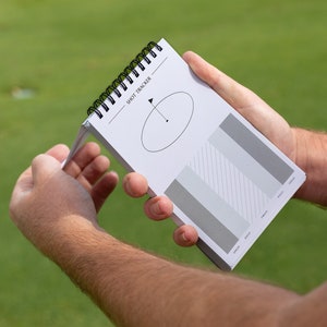May include: A white spiral bound notebook with the text "Shot Tracker" printed on the cover. The notebook has a diagram of a golf swing and a chart with grey bars.
