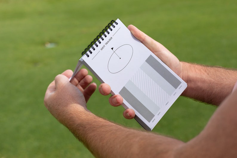 May include: A white spiral bound notebook with the text "Shot Tracker" printed on the cover. The notebook has a diagram of a golf swing and a chart with grey bars.