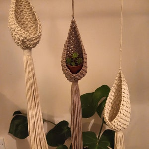 May include: Three macrame plant hangers in beige and grey tones are suspended from the ceiling. The hangers have a teardrop shape with a woven design and long tassels. One hanger holds a small potted plant with green leaves. The background includes large green leaves.