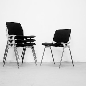 Puede incluir: Sillas negras y plateadas. Una silla está sola, mientras que las otras están apiladas. Las sillas tienen asientos y respaldos negros, con patas y marcos de metal plateado. El fondo es una pared blanca lisa.