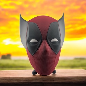 May include: A red and black Deadpool mask with a textured surface and gray ear-like extensions. The mask features black eye shapes and a mesh-like texture on the face. It is set against a blurred background of a sunset and a wooden table.