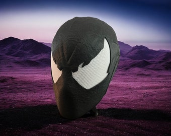 May include: A black superhero mask with white mesh eye openings. The mask is textured and appears to be made of a durable material. The background features a purple landscape with mountains and a blue sky.