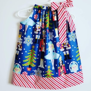 May include: A blue halter dress with a Christmas-themed print featuring nutcrackers, ballerinas, Christmas trees, and snowflakes. The dress has a red and white striped trim and a matching bow on the shoulder.