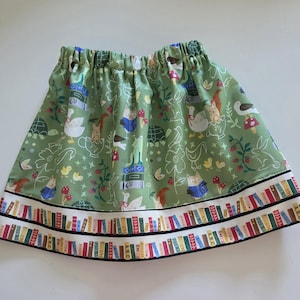 May include: A green skirt with an elastic waistband featuring a whimsical print of animals, mushrooms, and books. The hem is decorated with two horizontal bands displaying a colorful pattern of book spines with text including Poems and A.B.C.
