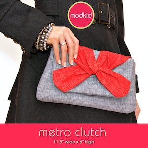 May include: A grey clutch bag with a large red bow. The clutch is 11.5 inches wide and 6 inches high. The text "metro clutch" is printed below the image.