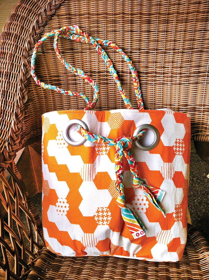May include: A white and orange tote bag with a geometric pattern. The bag has two grommets with a braided fabric strap going through them. The strap is made of different colored fabrics, including red, yellow, blue, and green.