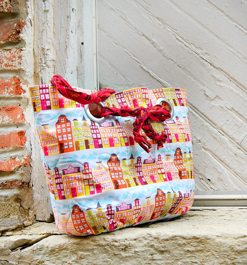 May include: A red and white quilted tote bag with a city skyline pattern. The bag has a red fabric handle with silver grommets.