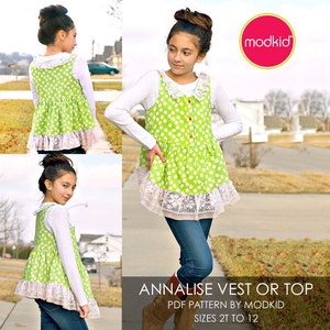 May include: A green and white polka dot vest or top with a white lace trim. The garment is worn over a white long-sleeved shirt with a white lace collar. The model is wearing blue jeans and brown boots. The text "ANNALISE VEST OR TOP" is at the bottom of the image. The text "PDF PATTERN BY MODKID" is below the first line of text. The text "SIZES 2T TO 12" is below the second line of text.