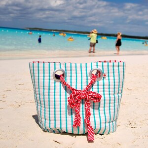 May include: A white and turquoise striped beach bag with red ribbon handles. The bag is sitting on a white sandy beach.
