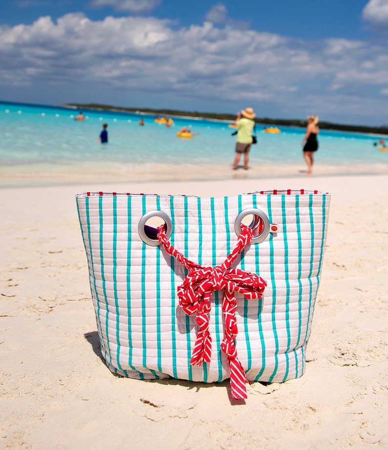 May include: A white and turquoise striped beach bag with red ribbon handles. The bag is sitting on a white sandy beach.