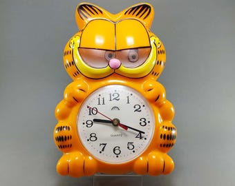 May include: A vintage orange Garfield-shaped quartz clock. The clock face is white with black numerals and hands. The clock has a pink nose and black stripes on the ears. The clock is a fun, decorative item.