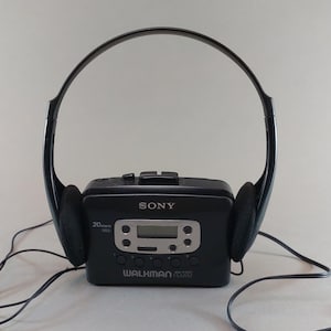 May include: A black Sony Walkman cassette player with attached black headphones. The Walkman features a digital display and control buttons. The headphones have black ear pads. The Walkman is connected to the headphones and a cable with a 3.5mm jack.