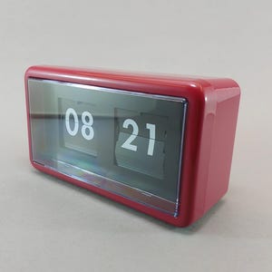May include: A rectangular flip clock with a glossy red plastic casing and a clear front panel. The internal display features black tiles with white numerals showing the time as 08 21. The clock has a smooth, rounded edge design.