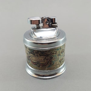 May include: A silver-toned lighter with a cylindrical body and a map design. The lighter has a hinged top and a small, round button. The map design features text that reads "ORBIS TERRARVM".