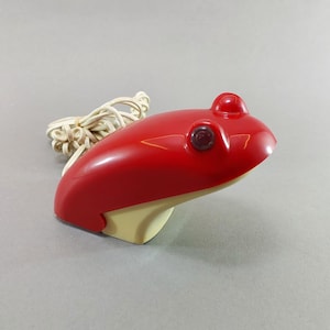 May include: A retro red frog-shaped desk lamp with a cream base. The lamp features a small red bulb and a round eye. A white cord is attached to the back of the lamp.