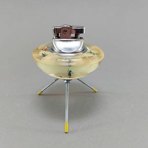 May include: A retro chrome lighter with a distinctive design. The lighter is mounted on a yellow, saucer-shaped base supported by three metal legs with yellow tips. The base has a painted landscape with small green figures.