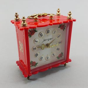 May include: A vintage red Jerger clock with gold accents. The clock features a white face with black Roman numerals and delicate rose illustrations. The clock has a gold handle and stands on small gold feet. The clock is approximately 4 inches tall.