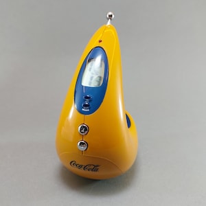 May include: A yellow Coca-Cola branded digital clock radio with a blue display and buttons. The clock radio has a unique teardrop shape, a silver antenna, and the Coca-Cola logo on the front.