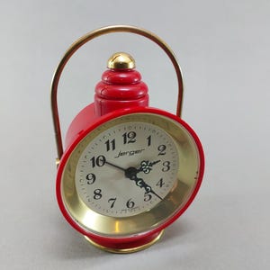 May include: A vintage red Jerger alarm clock with a gold-colored face and handle. The clock features a gold-colored base and a red, tiered top. The clock face displays black numerals and hands.