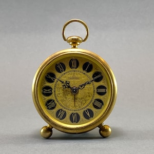 Peut inclure: Un réveil de couleur or avec des chiffres romains noirs et un motif floral sur le cadran. L'horloge indique "EUROPA 2 JEWELS".