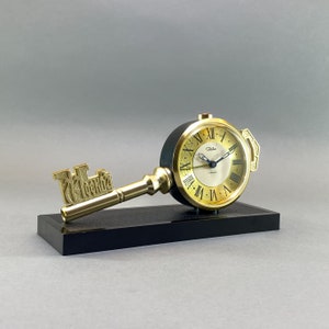 Può includere: Un orologio dorato a forma di chiave con una base nera. Il quadrante dell'orologio ha numeri romani e la parola "Tuba" stampata su di esso.