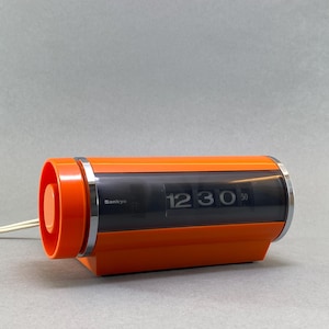 Könnte beinhalten: Eine orangefarbene und verchromte Sankyo-Klappuhr, die 12:30 Uhr anzeigt. Die Retro-Uhr hat ein zylindrisches Design mit einem dunkelgrauen Anzeigefenster und einem weißen Netzkabel. Die Uhrzeit wird in weißen Ziffern angezeigt.