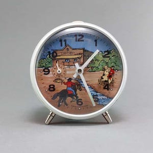 Könnte beinhalten: Ein weißer Vintage-Wecker mit einem Western-Thema. Das Zifferblatt zeigt einen Cowboy auf einem Pferd, der einen Stier fängt, eine Postkutsche und ein Gebäude mit dem Wort "peter". Die Uhr hat silberne Beine.