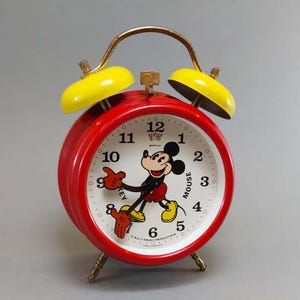 May include: A red Mickey Mouse alarm clock with yellow bells and a gold handle. The clock face features Mickey Mouse and black numerals. The clock has three brass-colored legs.