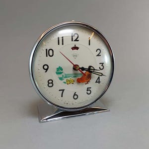 Può includere: Una sveglia vintage color argento con quadrante bianco. L'orologio presenta numeri neri, una lancetta dei secondi rossa e una piccola illustrazione di un edificio e un pollo. L'orologio è su un supporto argentato.