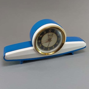 May include: A vintage blue and white clock with a gold-tone bezel and a cream-colored face. The clock has a rounded top and a rectangular base. The clock face displays black numerals and hands. The clock is a retro style.