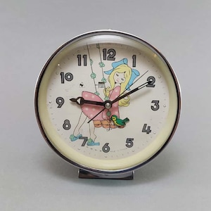 May include: A vintage, round alarm clock with a silver-tone metal frame. The clock face features a whimsical illustration of a girl on a swing, with a bird. The numbers are black, and the hands are black. The clock is approximately 4 inches in diameter.