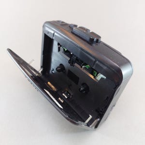 May include: A black portable cassette player with the cassette door open. The player has several buttons on top and a silver hinge. The interior reveals the mechanical components and circuit board.