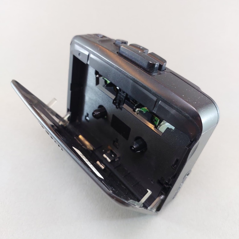May include: A black portable cassette player with the cassette door open. The player has several buttons on top and a silver hinge. The interior reveals the mechanical components and circuit board.
