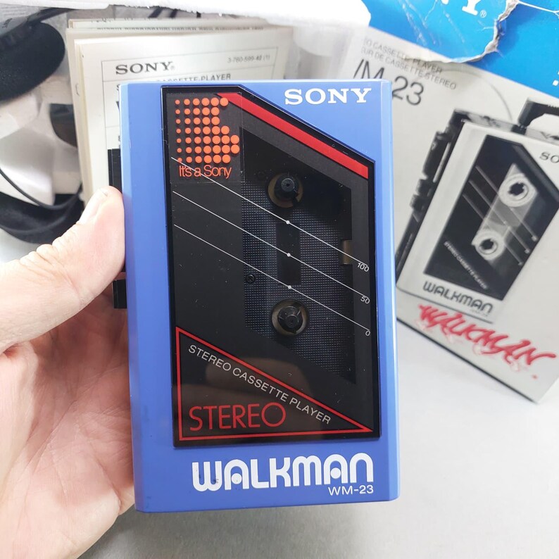 May include: A blue Sony Walkman WM-23 stereo cassette player featuring a black central panel with red accents, the text 'It's a Sony', 'STEREO CASSETTE PLAYER', 'STEREO', and 'WALKMAN WM-23'. The device includes a clear window showing the internal spools.