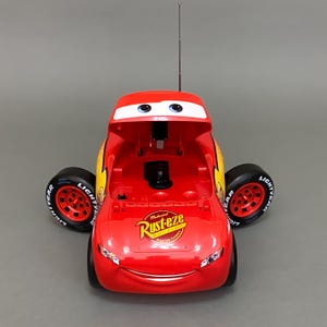 May include: Red toy car shaped like Lightning McQueen from the Disney Pixar Cars movie. The car has a cassette player with the hood open and a Rust-eze sticker on the side.
